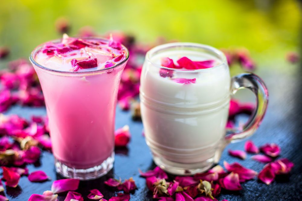 دو ماگ شیشه ای با نوشیدنی های خنگ سیز و صورتی رنگ که روی یک میز قرار داده شده و کنار آنها گلبرگ های گل رز صورتی قرار دارد