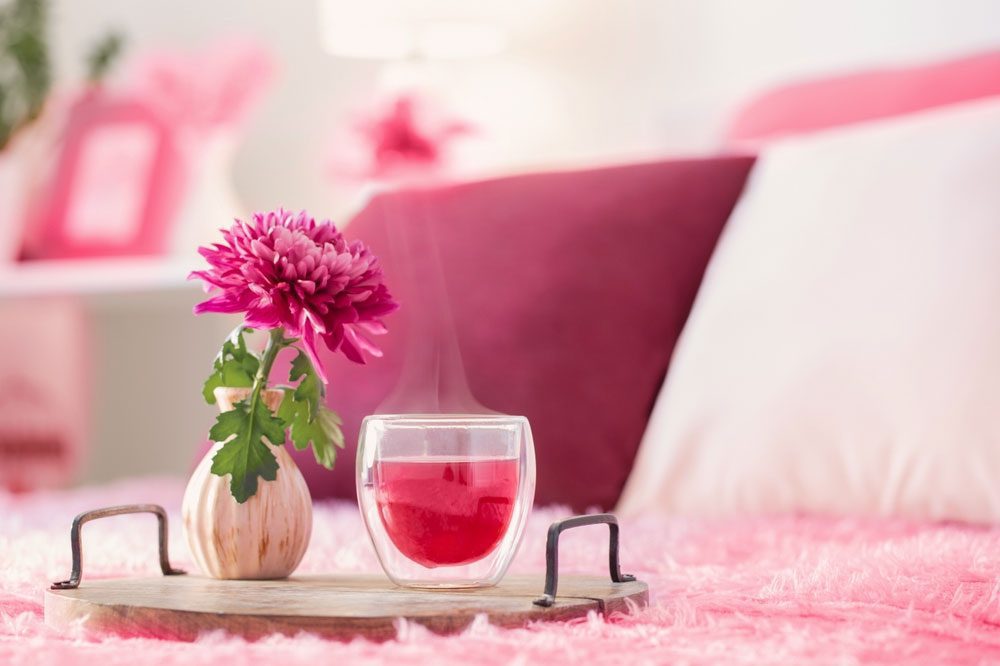 چای قرمز در یک ماگ شیشه ای دوجداره و عایق بندی شده در کنار یک گلدان و گلی زیبا روی یک رو تختی صورتی 
